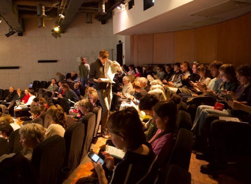 Zuhörer in einem Konferenzsaal; eine Person steht mit Unterlagen, Blick auf Bühne.
