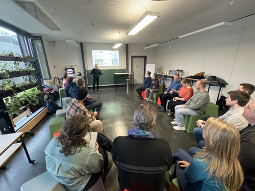 Gruppe von Menschen sitzt im Kreis in einem Seminarraum; Präsentation an der Wand
