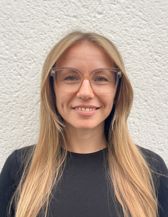 Porträt einer lächelnden Frau mit blonden Haaren und Brille vor hellgrauem Hintergrund.