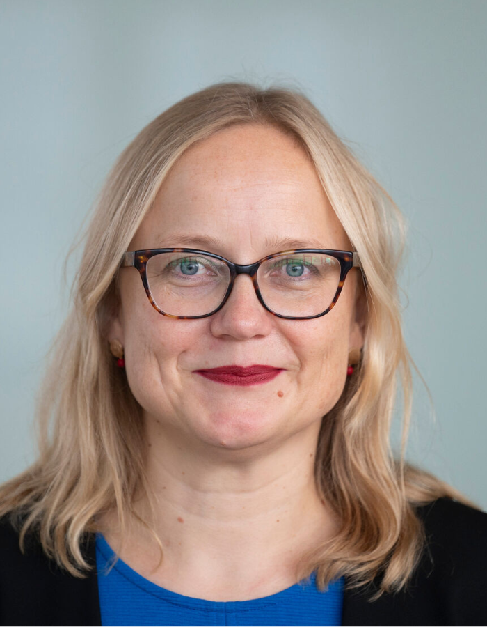 Melanie Arndt - Blonde Person mit mittellangen Haaren, blauem Oberteil und schwarzem Jackett vor grauem Hintergrund.