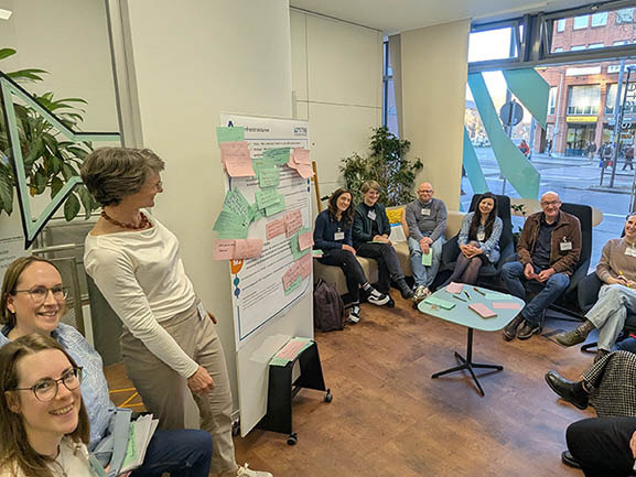 Gruppe sitzt in Kreis in Raum, Moderatorin steht bei Flipchart mit Post-its.