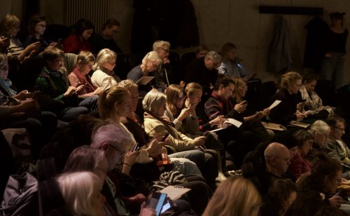 Publikum sitzt in einem Kino, zahlreiche Menschen schauen auf Fragebogen oder Geräte.