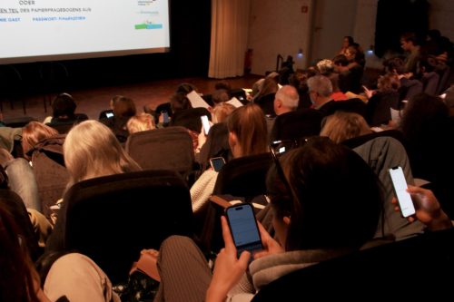 Personen in einem Konferenzsaal, viele schauen auf Smartphones und einen Präsentationsbildschirm.