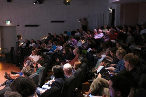 Publikum in einem Konferenzsaal, einige sitzen, andere stehen; Vortragende rechts spricht.