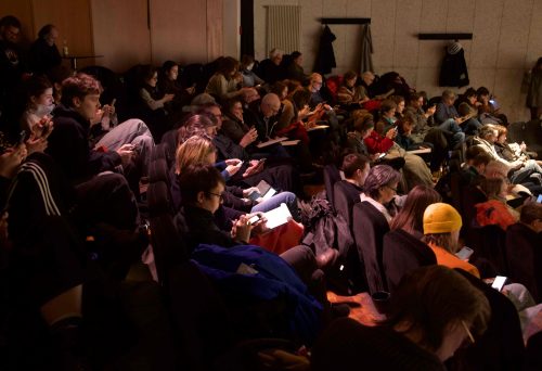 Versammelte Menschen in einem Saal, einige lesen Umfragebögen oder richten Blick nach vorne.